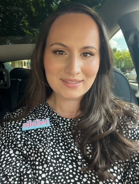 A woman with long dark hair smiling while sitting in a car, wearing a black and white polka dot top with a pink and blue name badge that says 'Malia'.