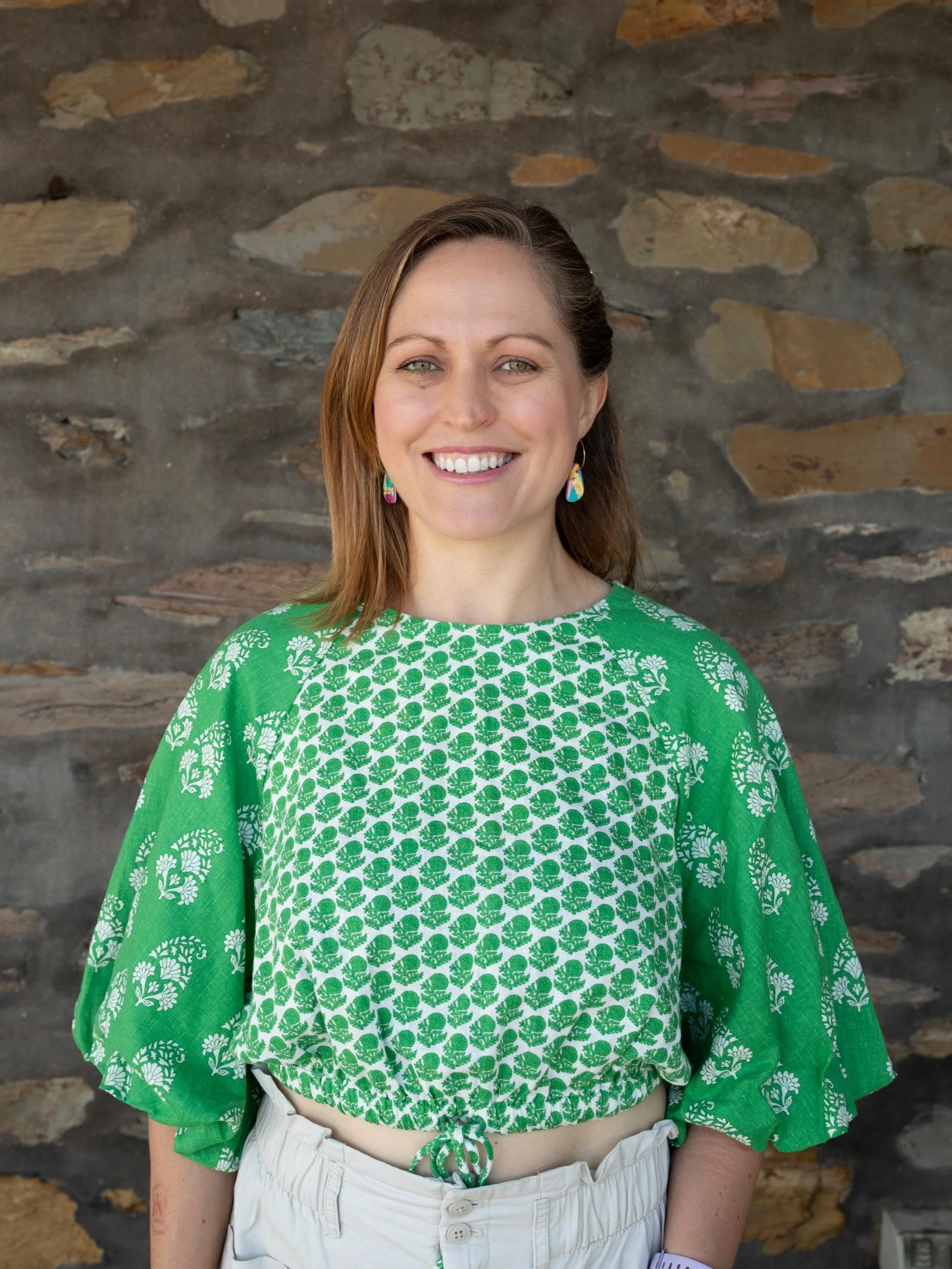A woman with brown hair and green eyes smiling, wearing a green patterned top and white high-waisted pants, standing in front of a stone wall.