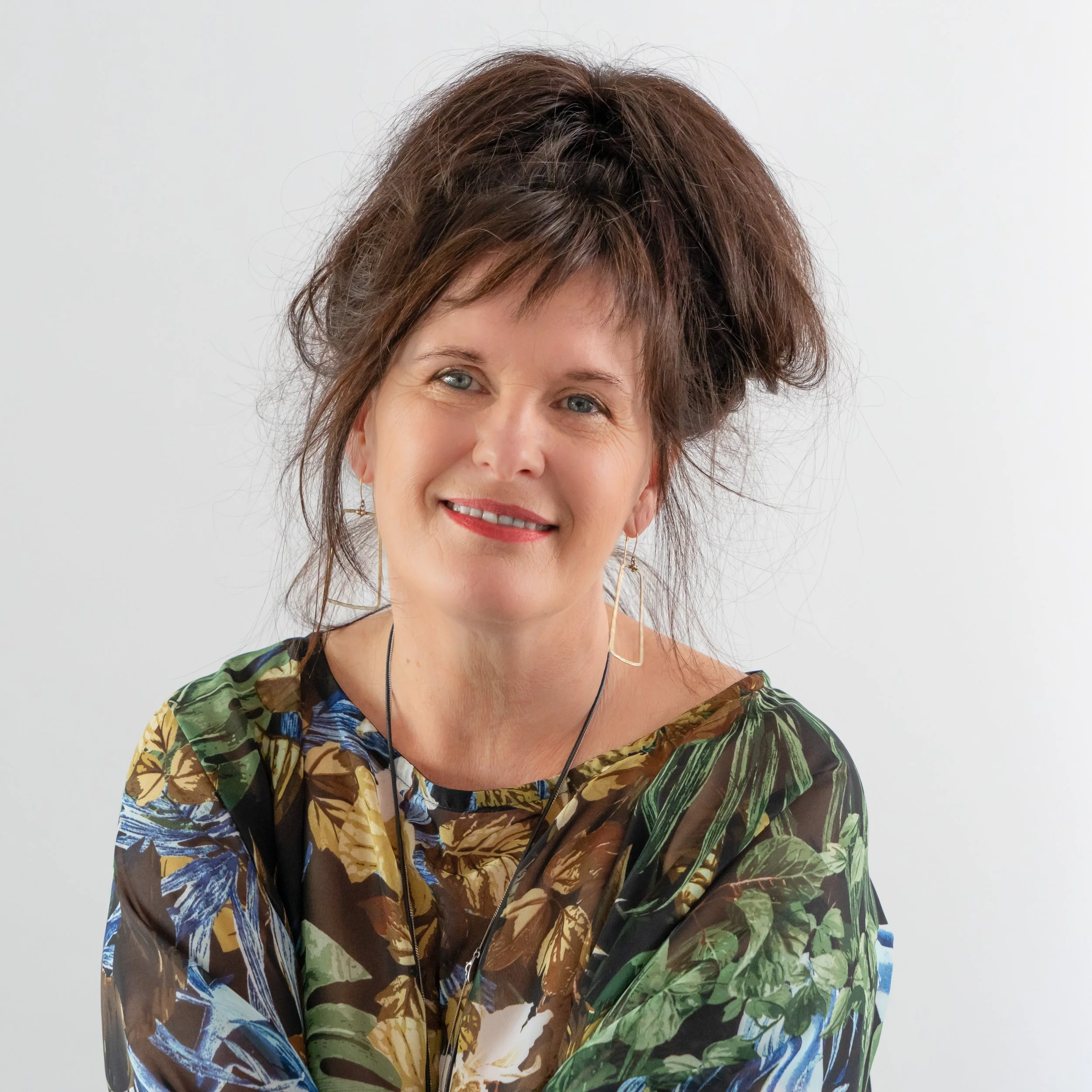 Close-up of a woman with short, layered brown hair, wearing colorful patterned clothing with green, blue, and brown leaves, smiling softly against a plain white background.
