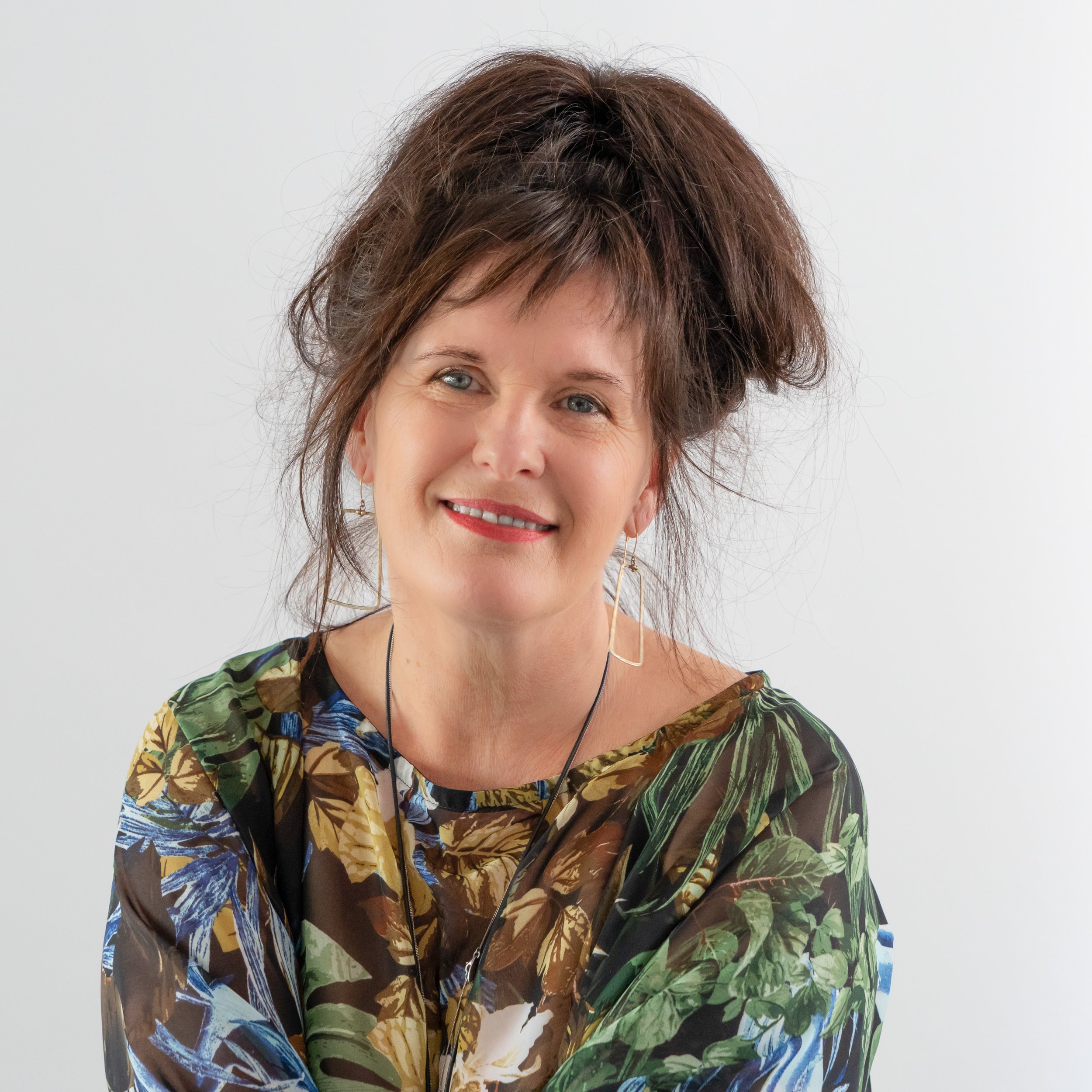A woman with short brown hair, blue eyes, and freckles, smiling against a white background. She is wearing a colorful, patterned top with green, brown, blue, and beige tones and large gold earrings.