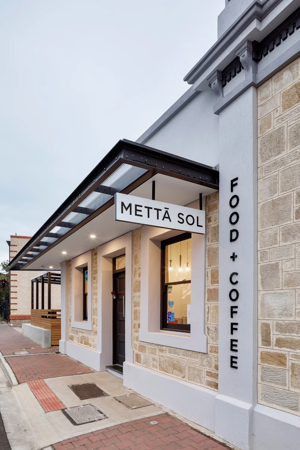 Exterior of a café with signs reading "Mettā Sol" and "Food + Coffee" on the building. The building has stone walls, a black door, and large windows, with a sidewalk in front.