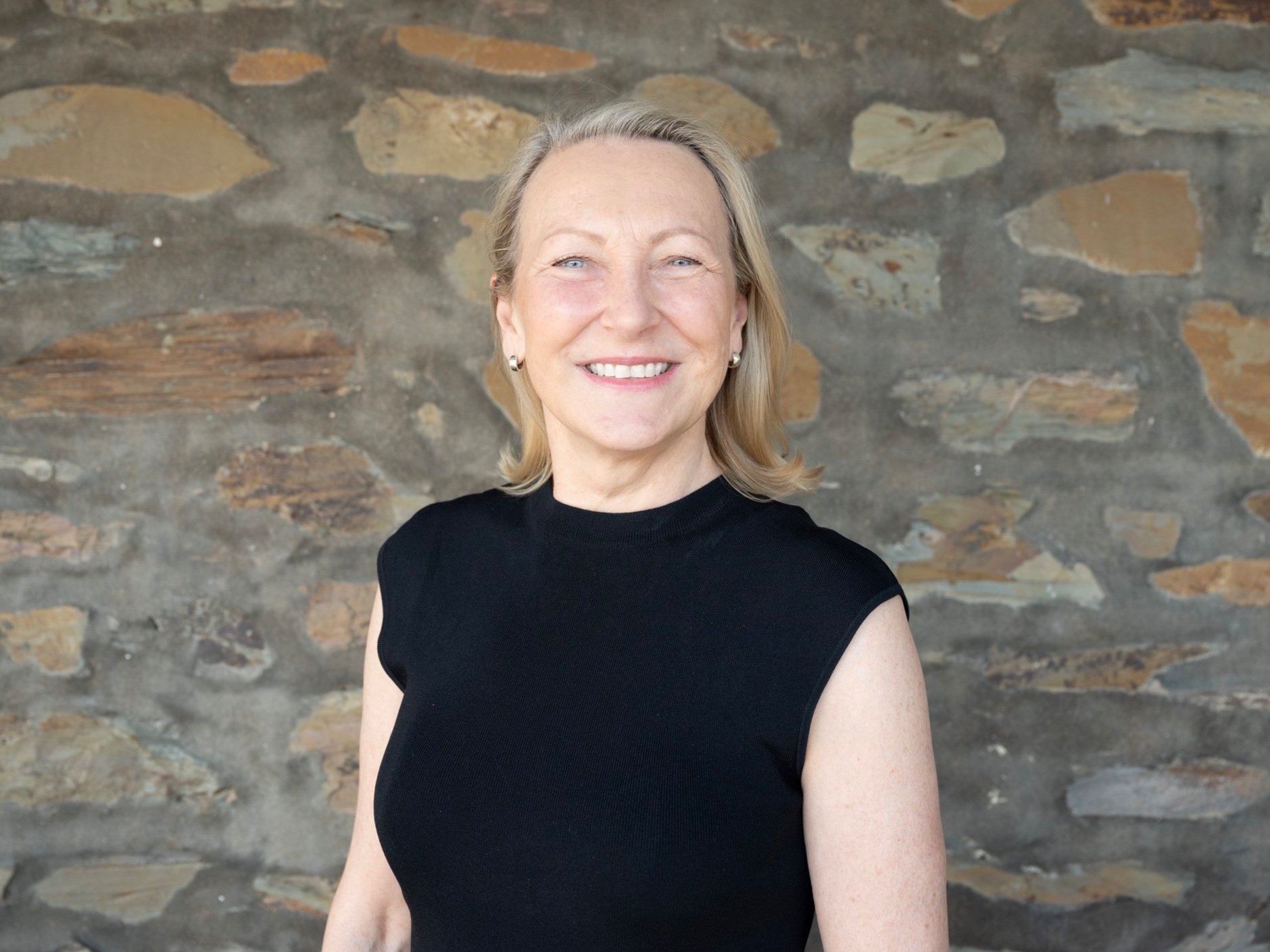 Portrait of a middle-aged woman with blonde hair, blue eyes, wearing a sleeveless black top and small earrings, smiling, standing in front of a stone wall.