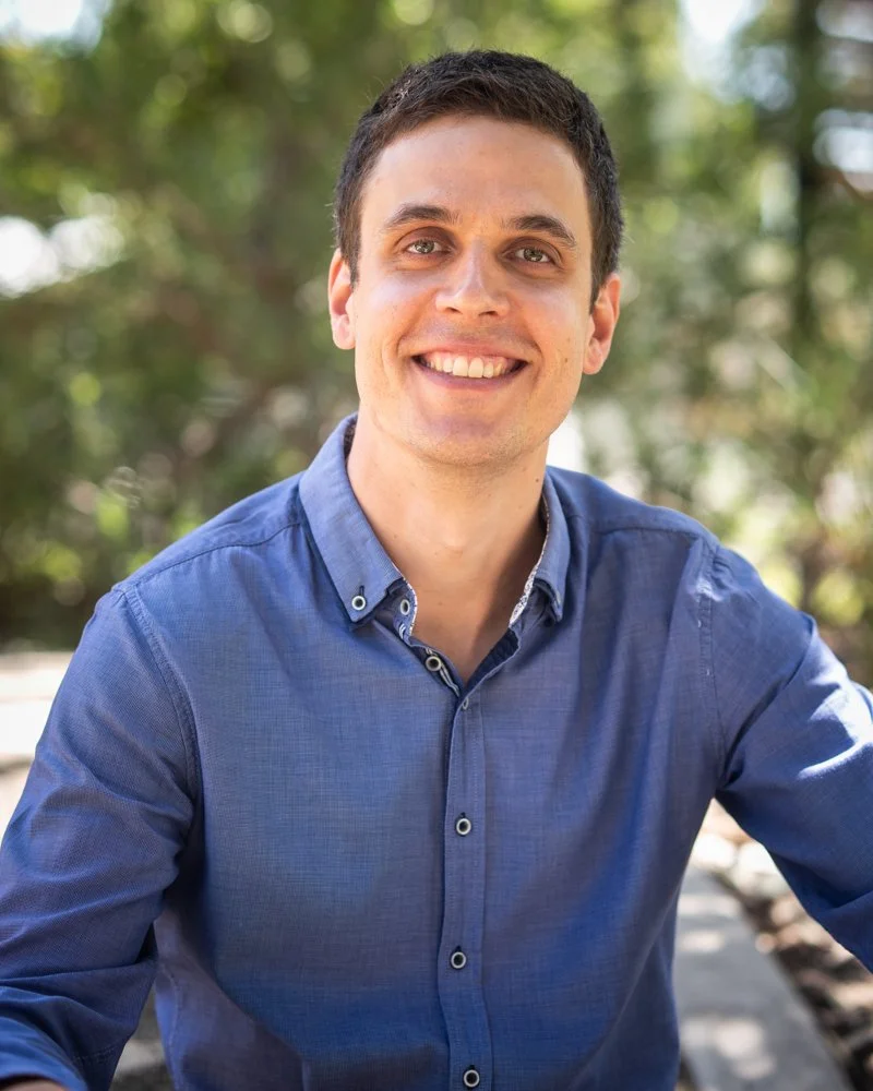 Smiling man wearing a blue button-up shirt outdoors with blurred green trees in the background.