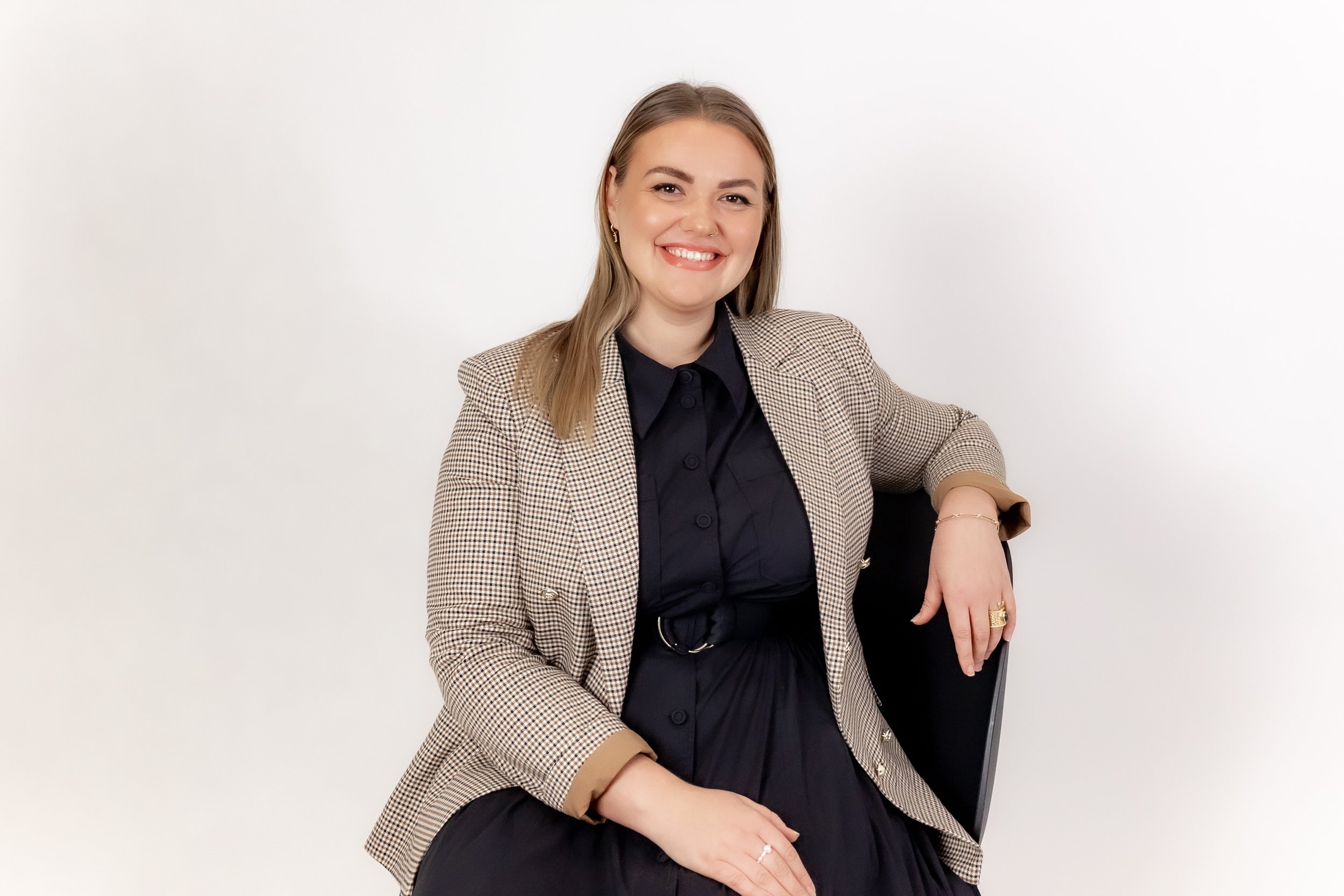Young woman with light brown hair smiling, wearing a checkered blazer and black dress, sitting on a black chair against a plain white background.