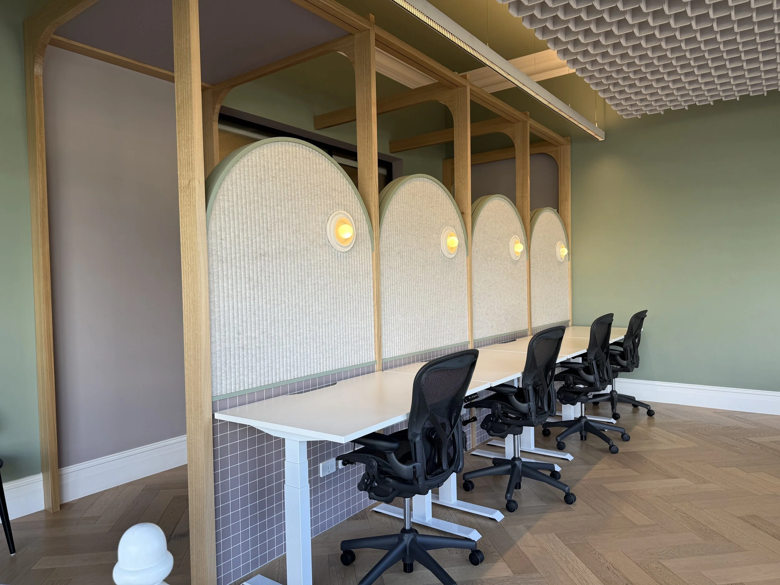A modern office space with a long white desk, ergonomic black chairs, and a decorative partition with a wavy design, small circular lights, and a wooden frame. The room has a green wall, a ceiling with acoustic panels, and a wood floor.