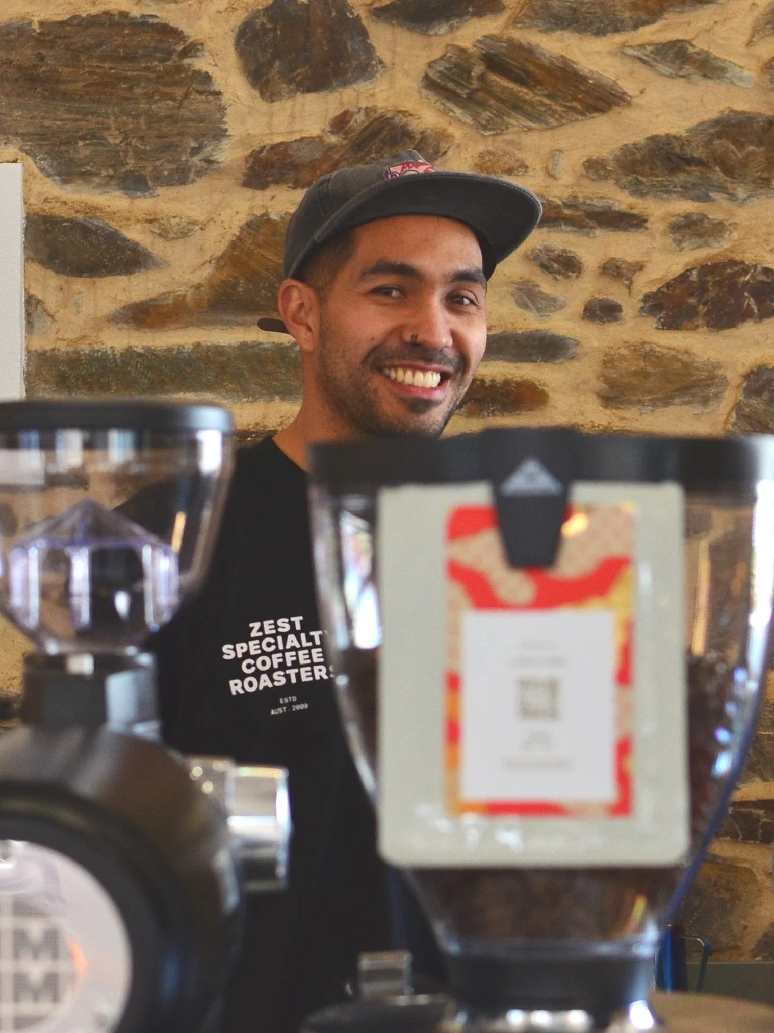A young man with a cap smiling at the camera, behind a coffee bag labeled "st zest specialty coffee roaster," with a stone wall in the background.