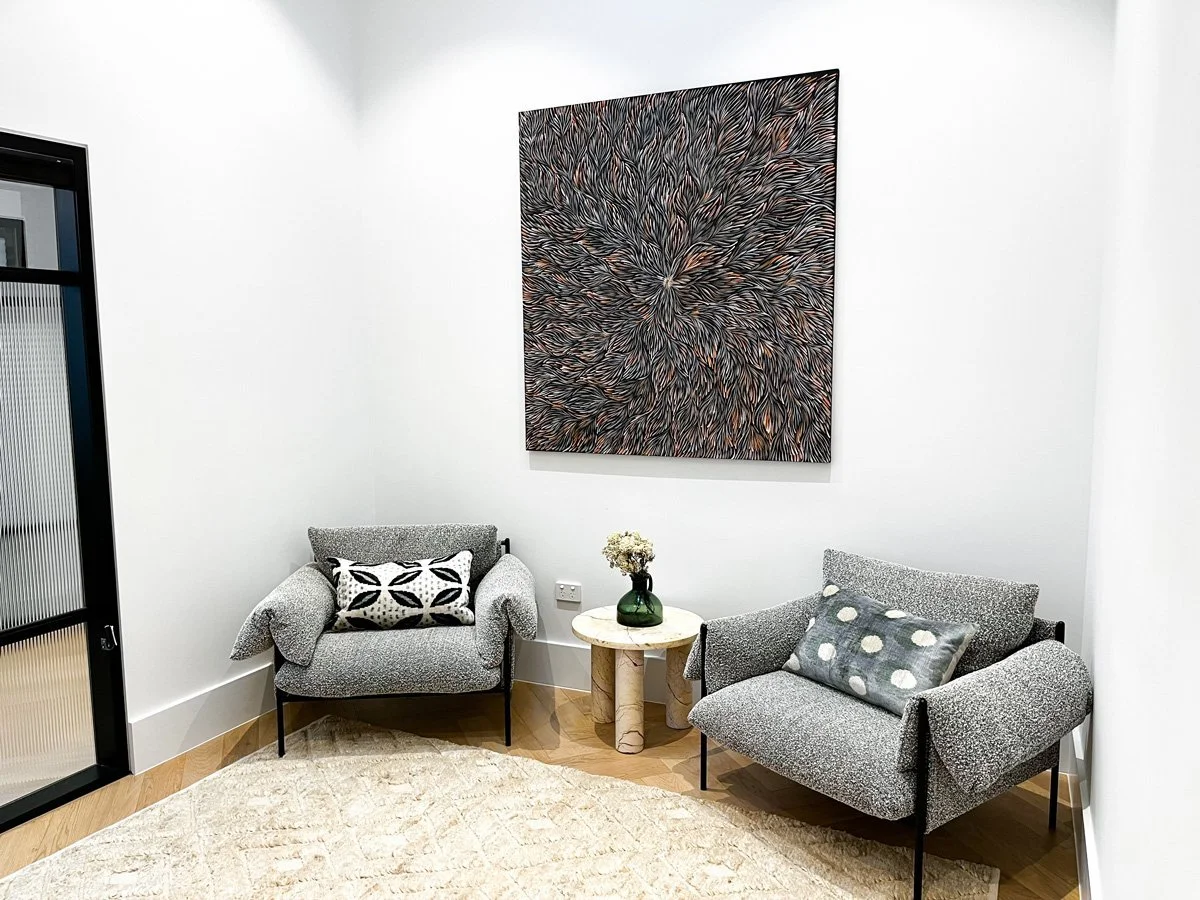 Modern living room corner with two gray armchairs, a small round marble side table with a green vase and white flowers, a large abstract artwork on the white wall, a cream-colored rug, and a sliding glass door.