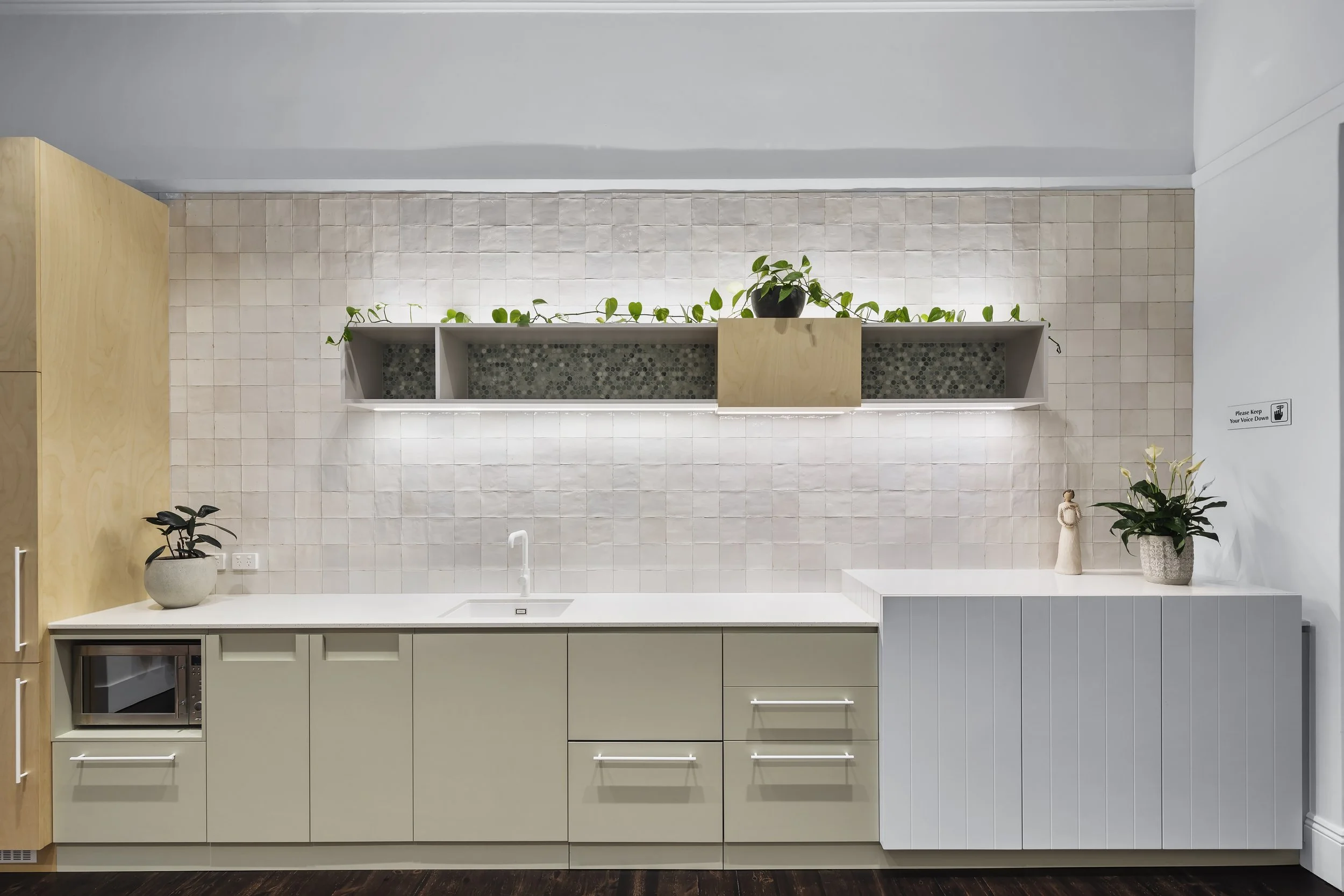 A modern kitchen with beige cabinets, a white countertop with a sink, and a backsplash of beige tiles. There are plants on top of the cabinets and on the counter, a microwave, and a small decorative figure near the right side.