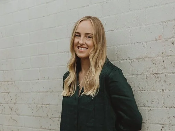 A woman with long blonde hair and a black jacket smiling, standing against a white brick wall.