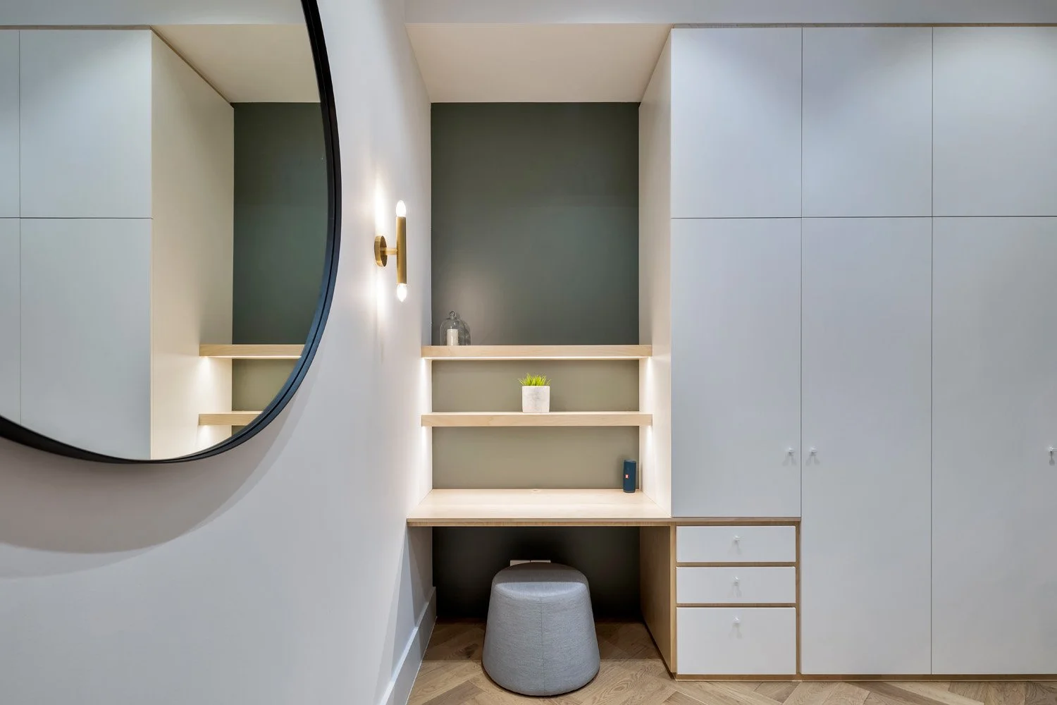 Modern corner of a bedroom or dressing room with large white wardrobe, built-in desk with small drawers, and a gray ottoman. A round mirror hangs on the wall and a wall sconce provides lighting. Decorative items on shelves include a small plant and a glass container.