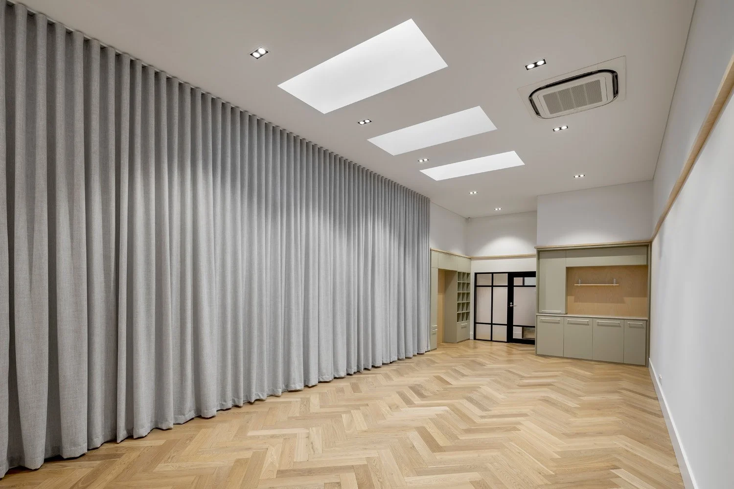 An empty room with a wooden herringbone floor, beige cabinetry, a white wall, a black framed door, ceiling skylights, and an air conditioning unit.