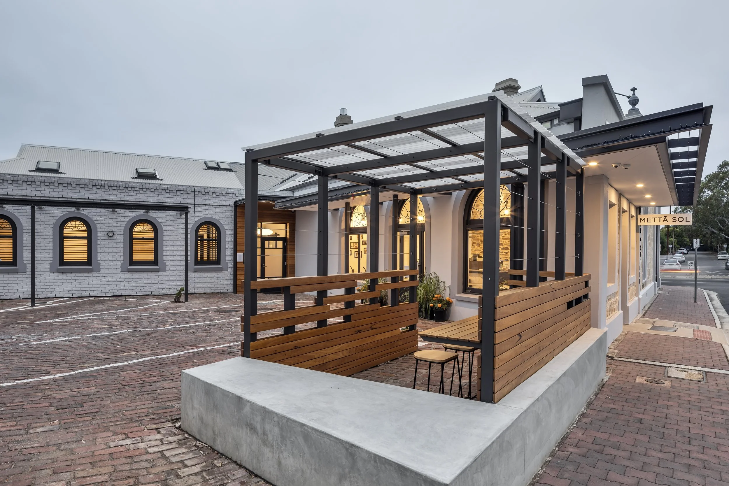 A modern building with a wooden and metal pergola, illuminated windows, and a sign that reads 'METTÄ SOL' in front of a brick sidewalk.