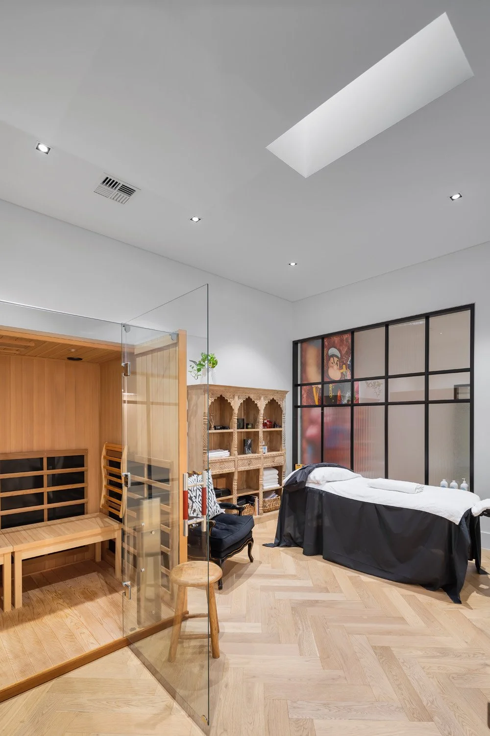 A spa treatment room with a massage table covered with white sheets and a black skirt, wooden shelves with towels and decor, a sauna with a glass door, and a large window with colorful artwork.