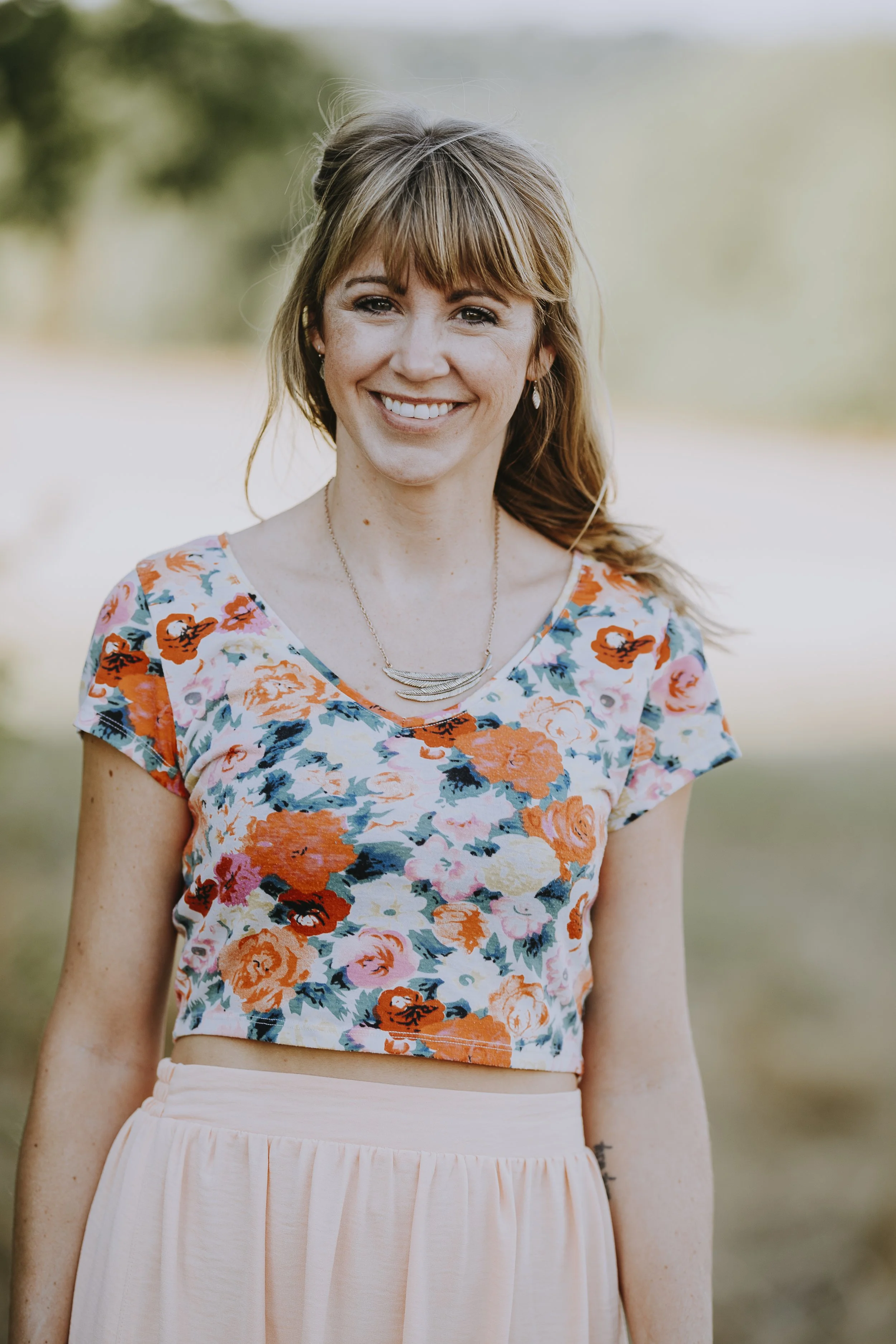 A woman with light skin, shoulder-length light brown hair, and a smile, standing outdoors on a sunny day. She is wearing a colorful floral crop top, a light pink high-waisted skirt, a delicate necklace, and earrings. The background is blurred with green foliage.