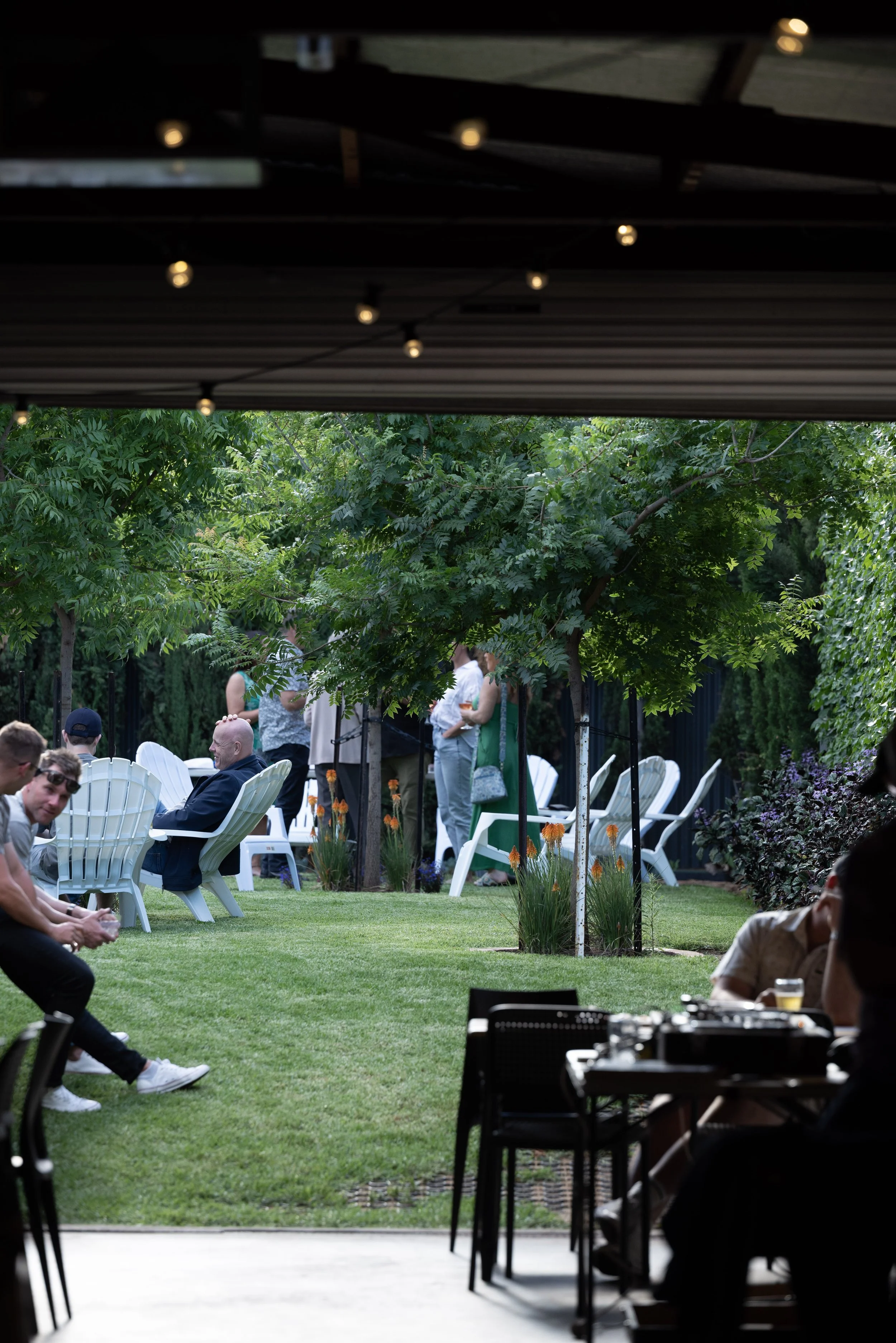 People gathered outdoors at a garden party or event, sitting on white chairs and standing among green trees and plants, with some wearing sunglasses and casual clothing, under a dark roof with string lights.