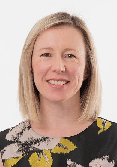 A woman with blonde hair smiling, wearing a black top with yellow and white floral patterns, against a white background.