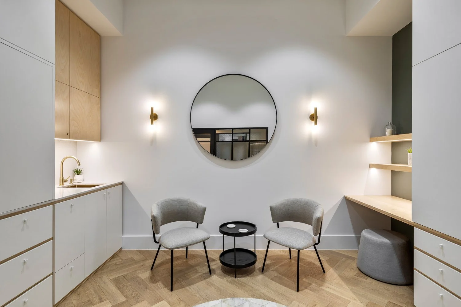 Minimalist interior with two grey chairs, a small black round table, a large circular mirror on a white wall, two wall-mounted gold light fixtures, white and wooden cabinetry, and built-in shelves with decorative items.