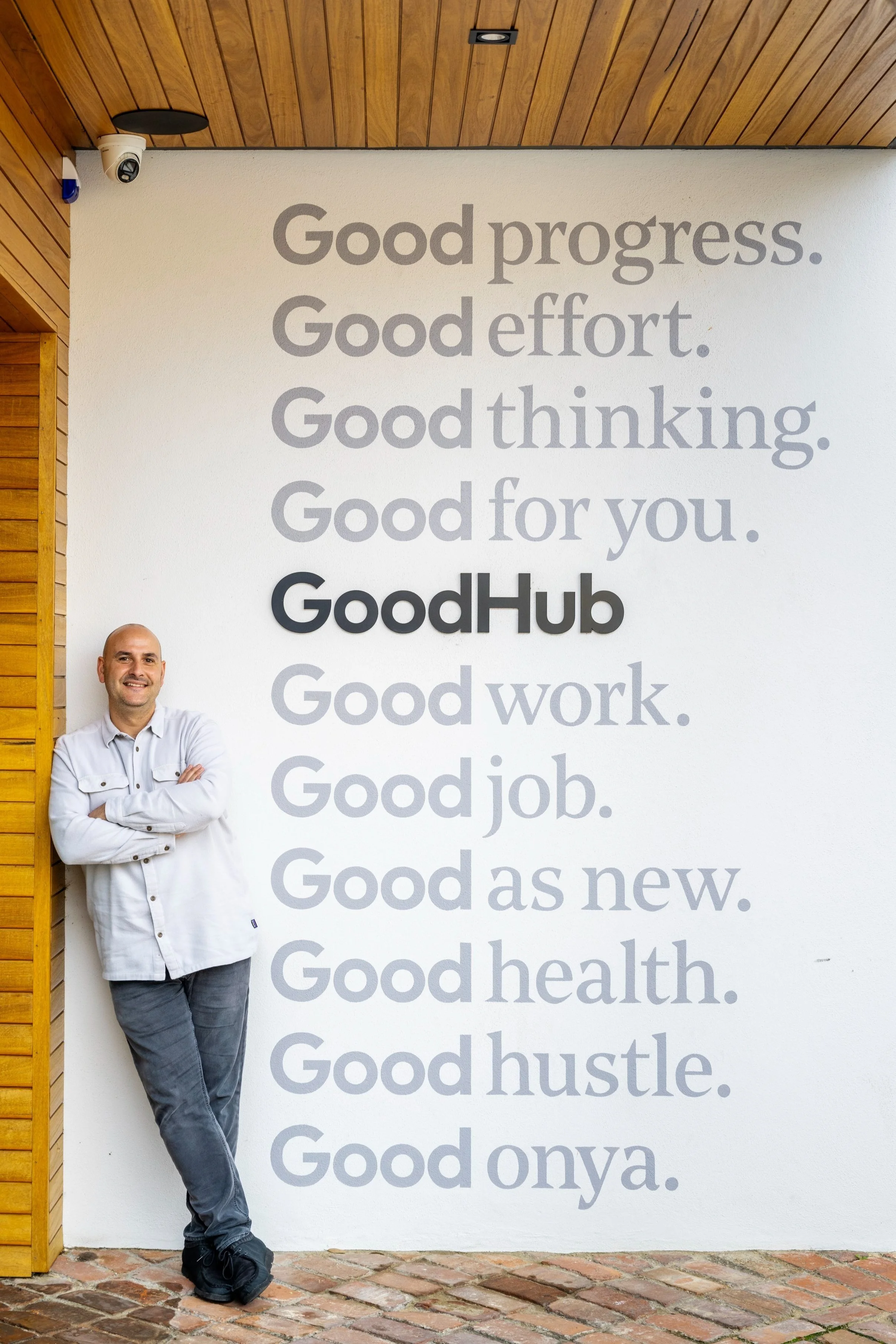 A smiling man stands with arms crossed in front of a wall with a motivational quote. The wall has wooden panels on the ceiling and some security cameras in the upper corners. The quote promotes positive attributes such as good progress, effort, thinking, and work, ending with good health and hustle.