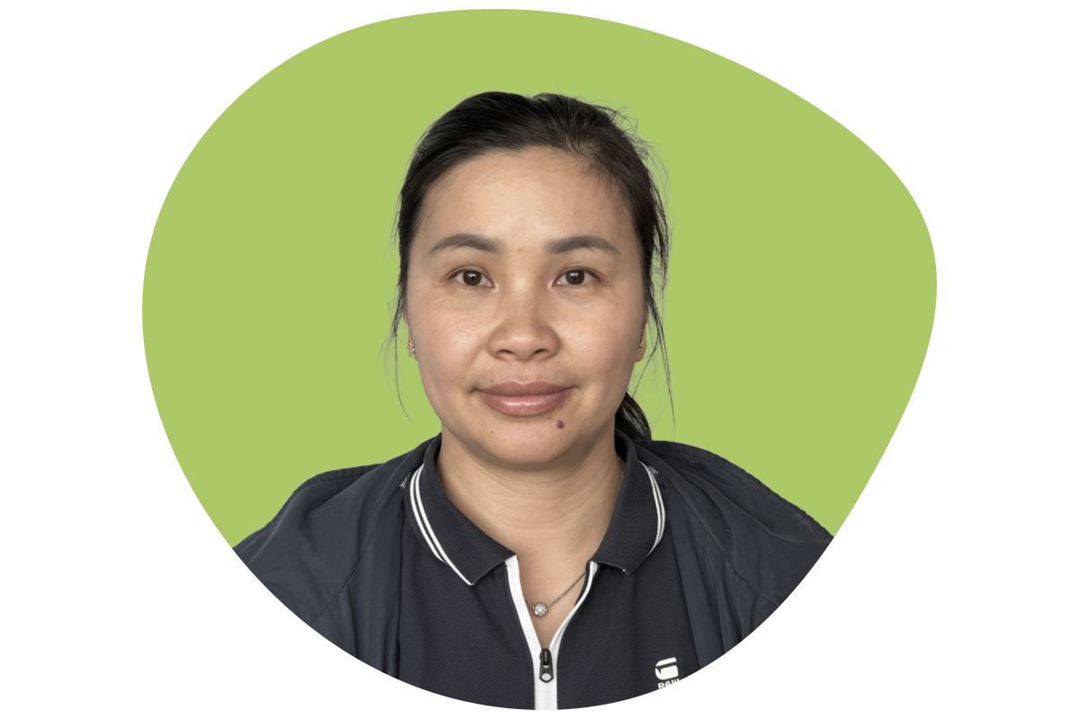 A woman with long brown hair smiling, wearing a black shirt and a silver necklace with a cross, against a background with black and green sections.