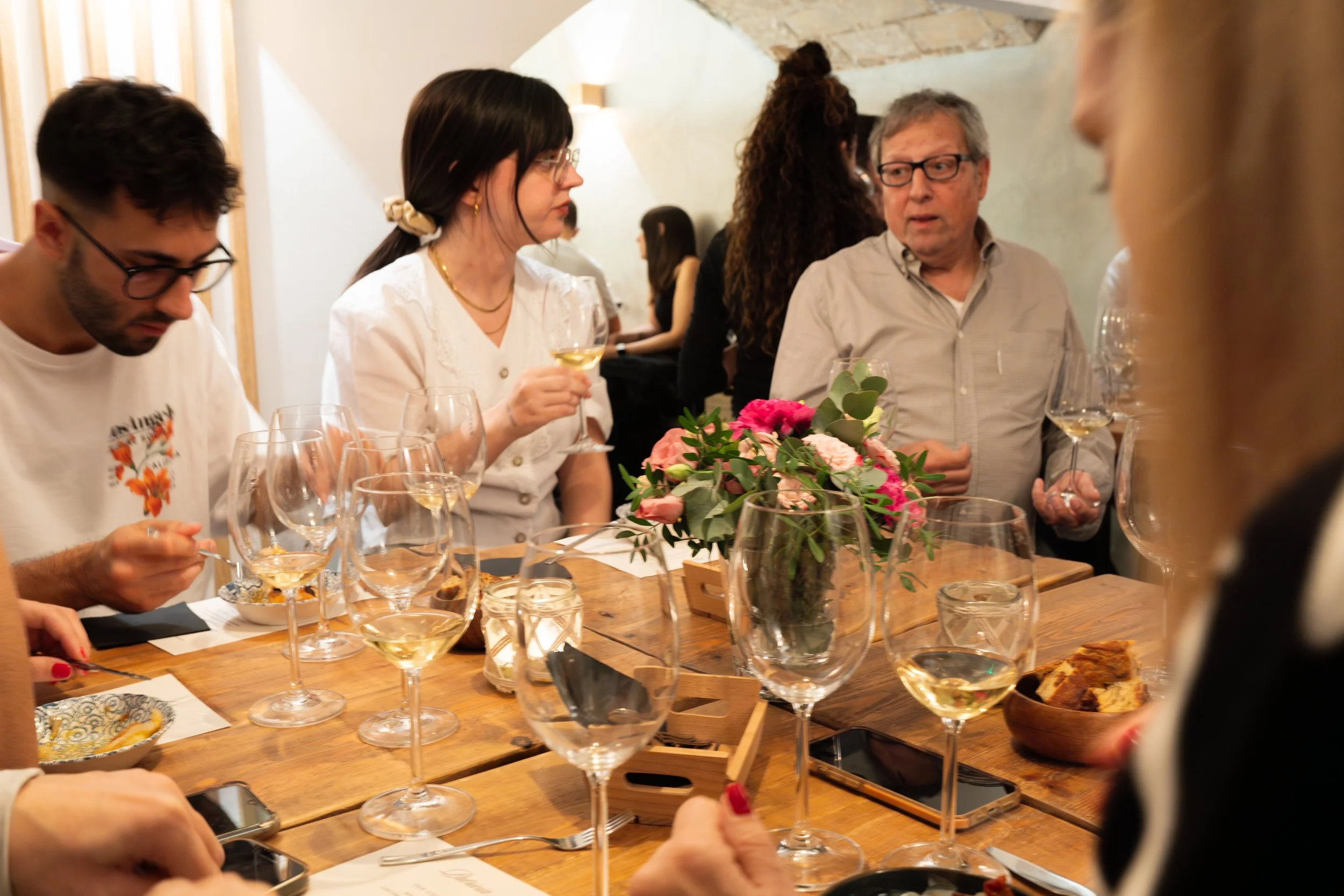 Varias personas en una mesa con copas de vino, una florero con rosas rosas y verdes, y comida, en un ambiente de comedor.