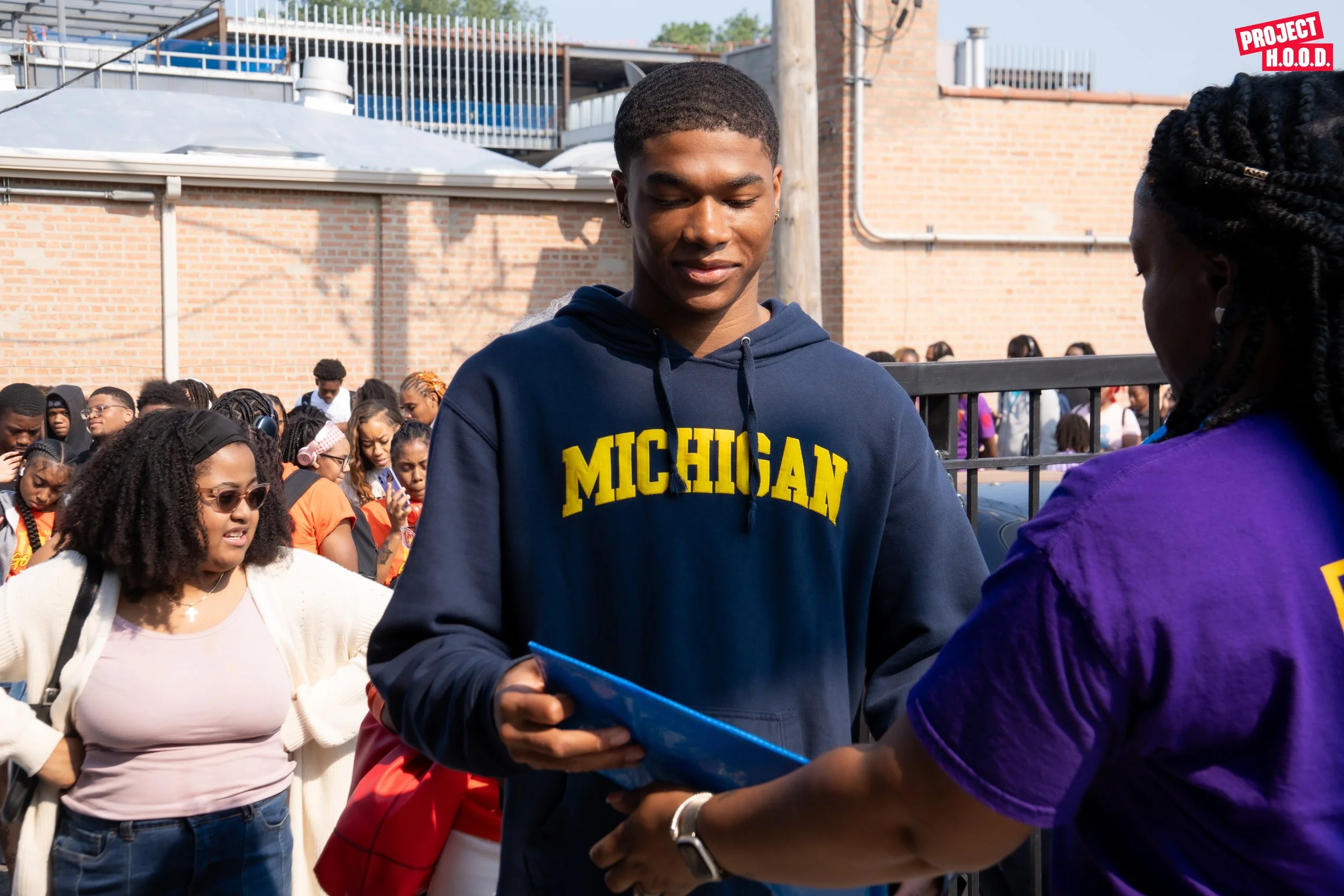 A young man wearing a Michigan hoodie is receiving a document or certificate from a woman in a purple shirt outdoors during a crowd event.