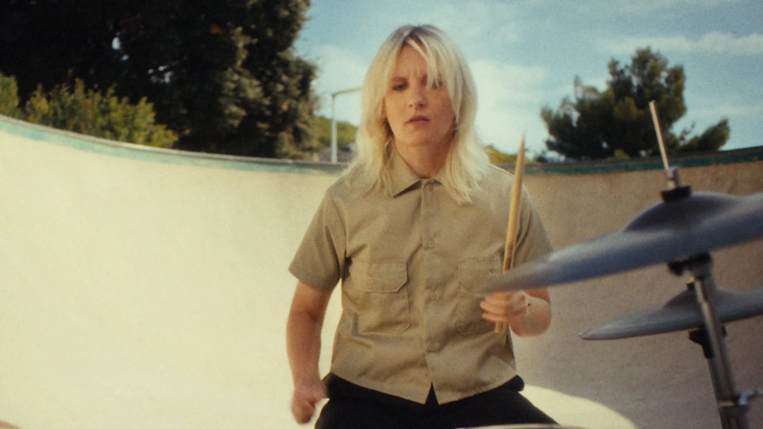 Marion Brunetto from the band Requin Chagrin with blonde hair playing drums outdoors, holding drumsticks, with a yellow wall and trees in the background.