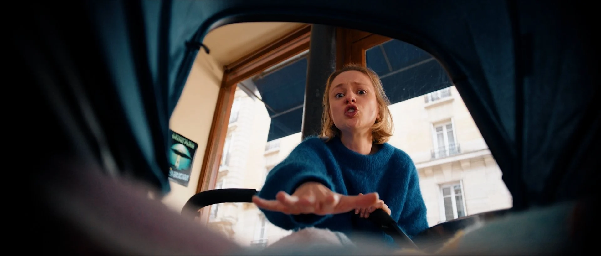 Tiphaine Daviot with brown hair wearing a blue sweater appears to be speaking or yelling, seen from an unusual low angle, with a building and balcony in the background.