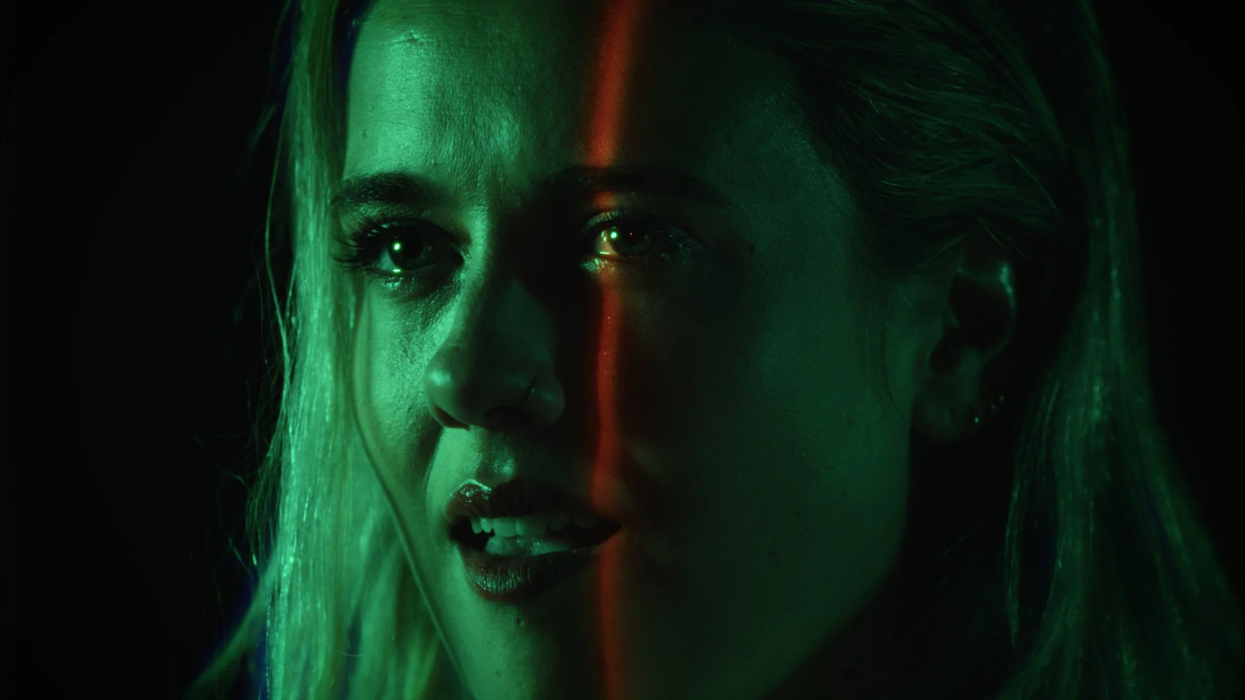 Close-up of a woman with blonde hair illuminated by green and red neon lights, with a dark background.