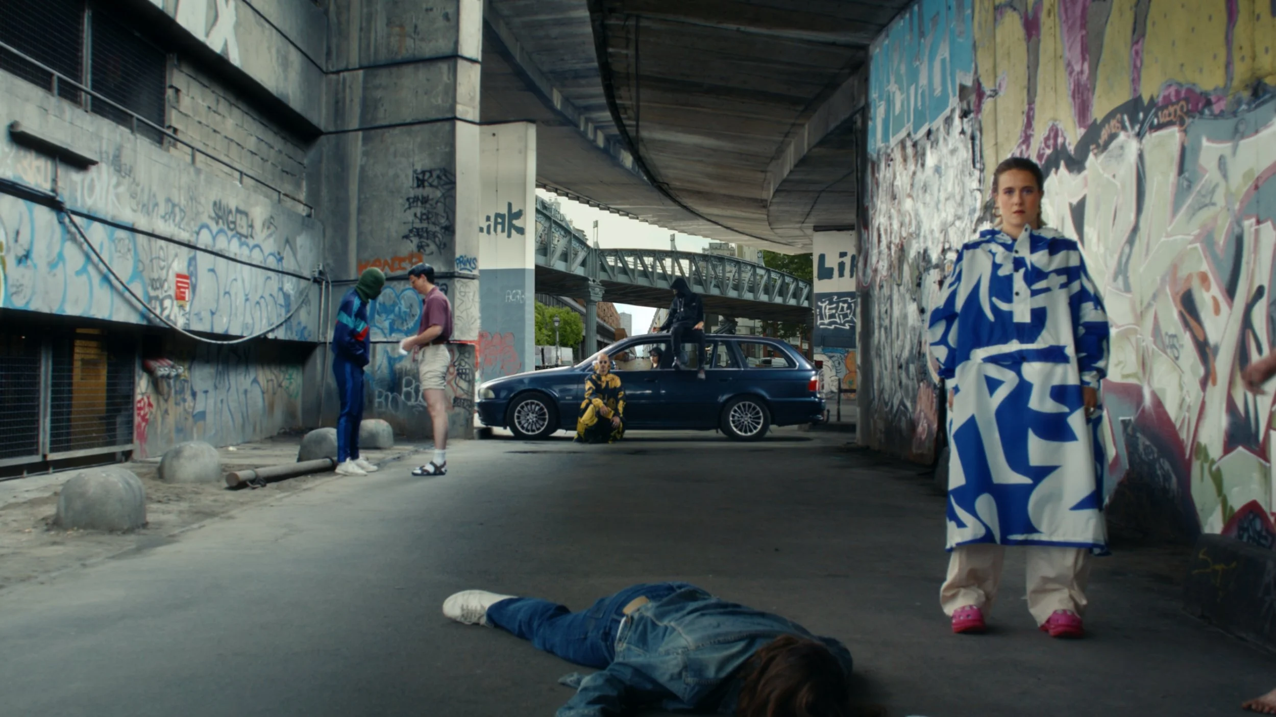 People under a graffiti-covered bridge, including a woman in a blue and white patterned coat standing next to a person lying on the ground, and others near a parked car, with an urban environment in the background.