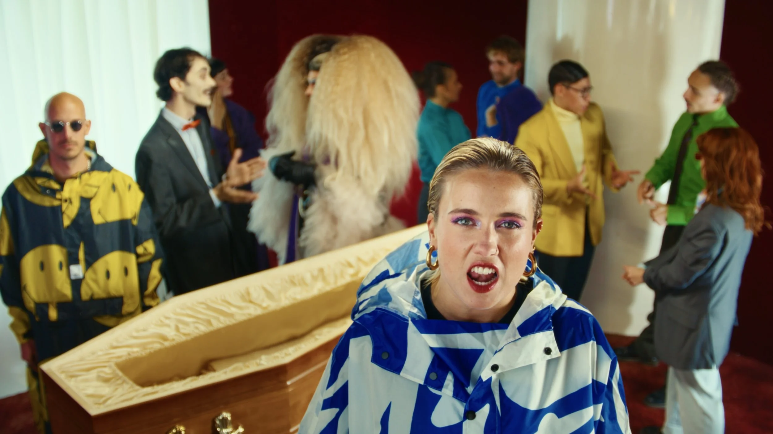A woman with blonde hair and hoop earrings appears angry or shouting in the foreground. Behind her, a diverse group of people are engaged in conversations, with some wearing colorful clothing, and a large fish-shaped coffin is visible on a table.