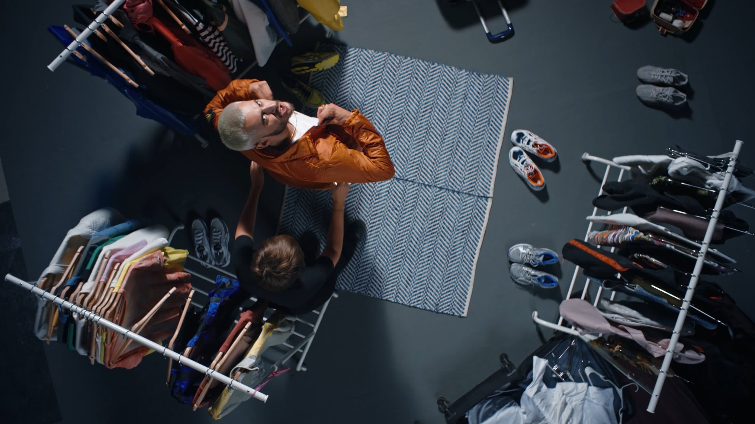 Overhead view of a man trying on an orange jacket in a cluttered room with clothes racks and shoes.