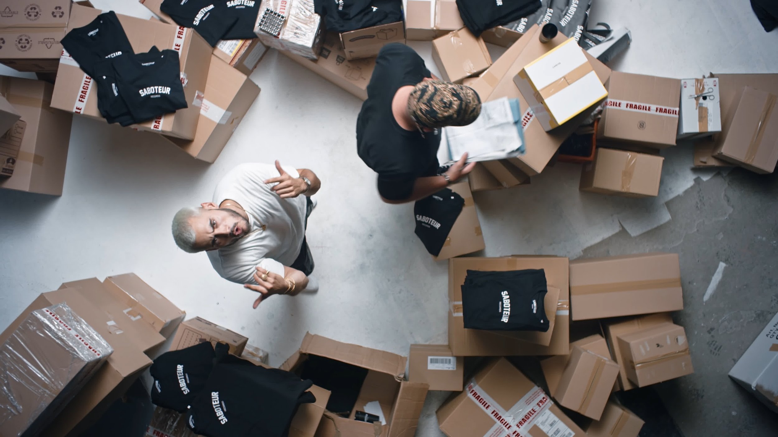 Two people surrounded by cardboard boxes and black t-shirts with "Saboteur" printed on them, viewed from above.