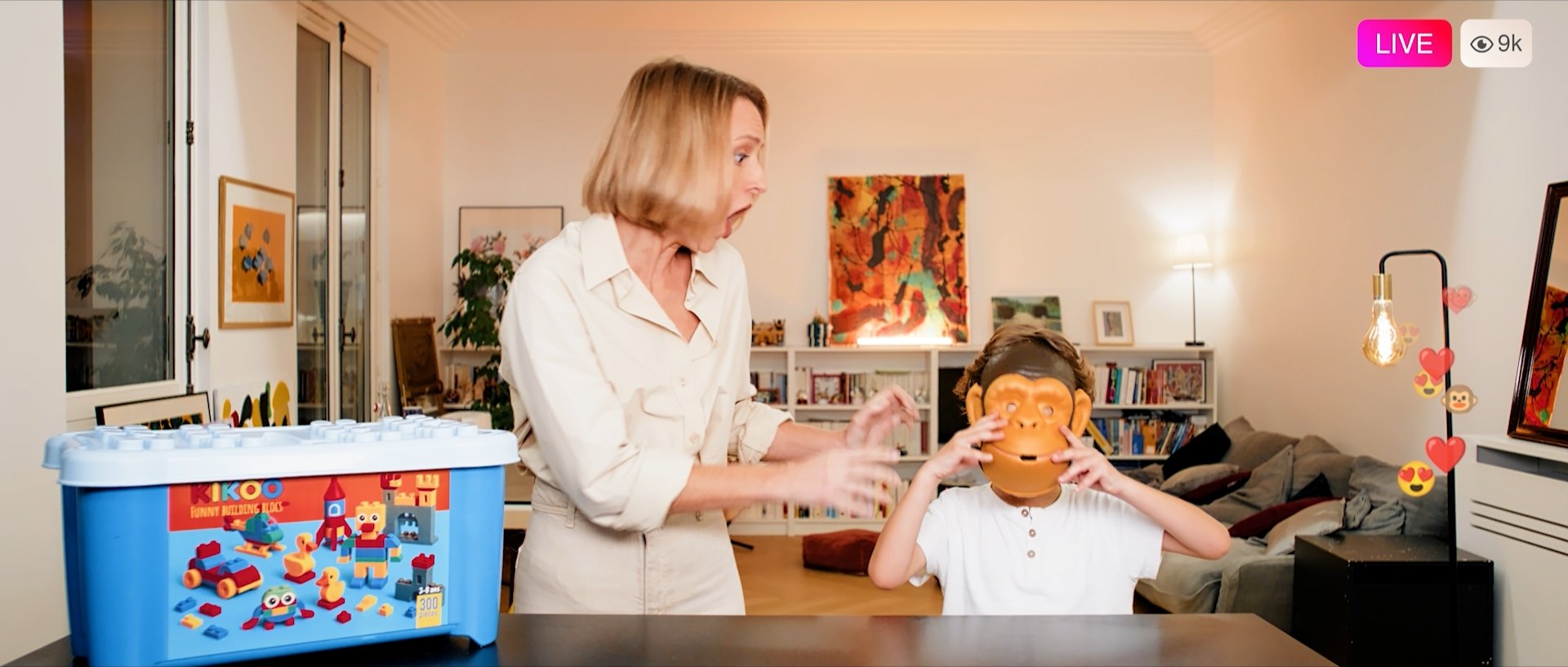 A woman helps a child put on a monkey mask in a cozy living room decorated with colorful artwork and books. The scene is well-lit and lively, with a toy building box on the table and lively hearts and love icons on the right side of the image. The image is being streamed live with 9,000 viewers.