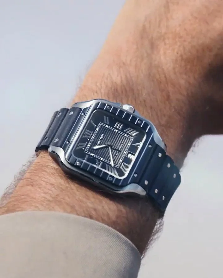 Close-up of a man's wrist wearing a Santos black and gray Cartier watch with a square face and Roman numeral markers.
