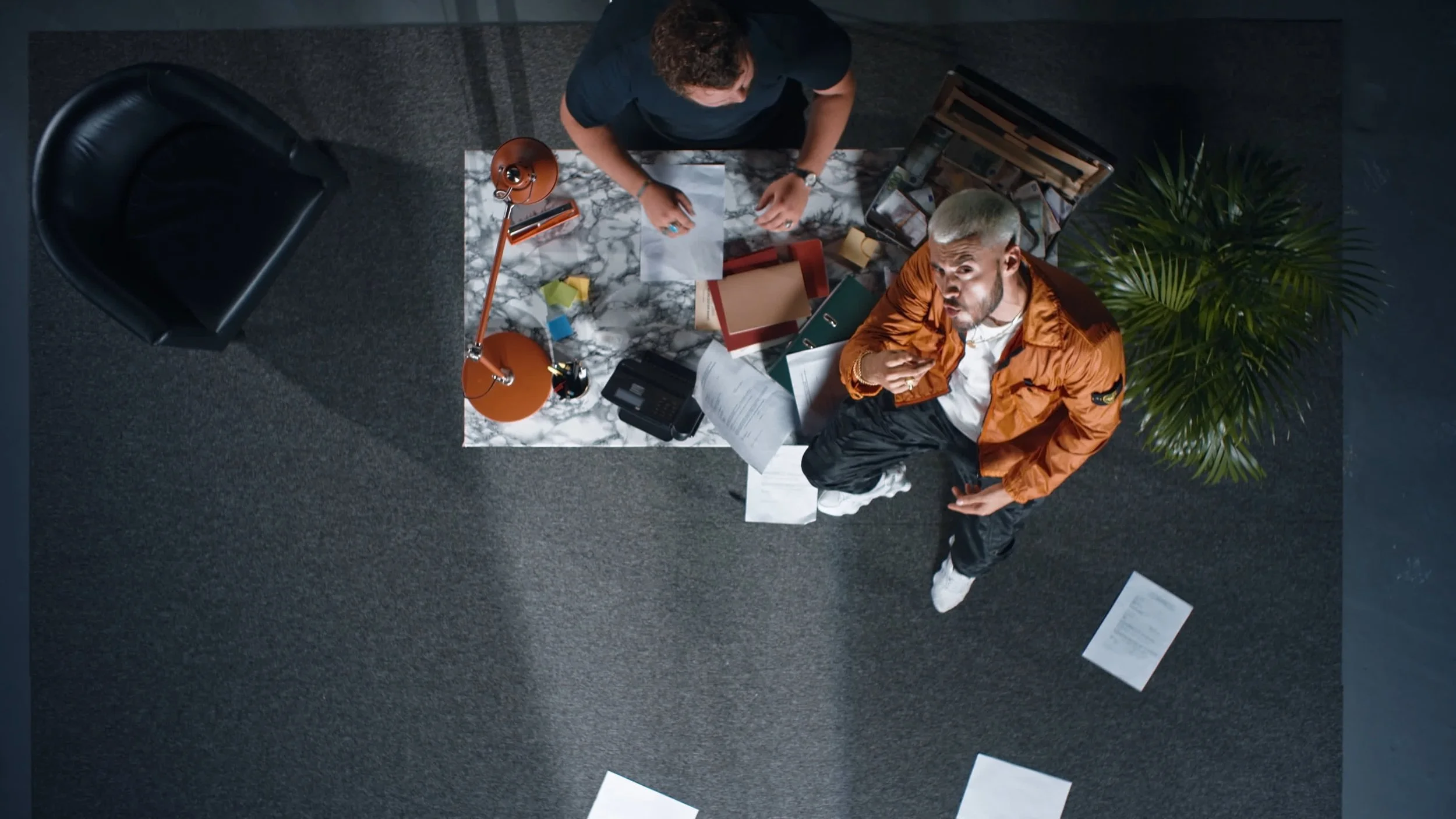 Overhead view of two people in an office setting, with a cluttered marble table, papers, a lamp, and greenery.