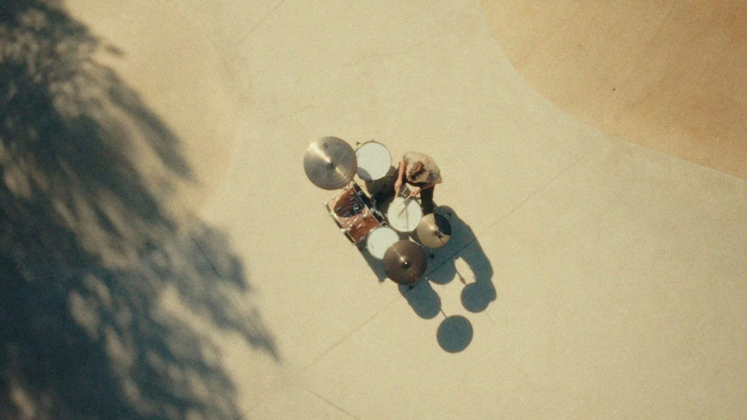 Top-down view of a Marion Brunetto from the band Requin Chagrin playing a drum set outside, with a tree casting a shadow on the ground.