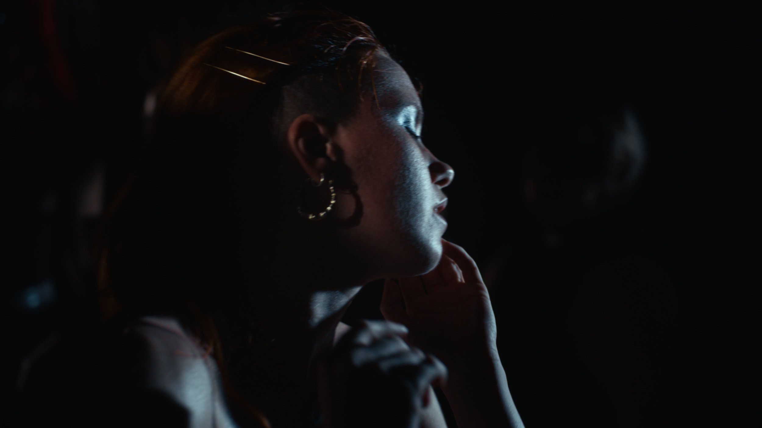 Side profile of a woman with earrings and braided hair, illuminated softly against a dark background, with her hand touching her chin.