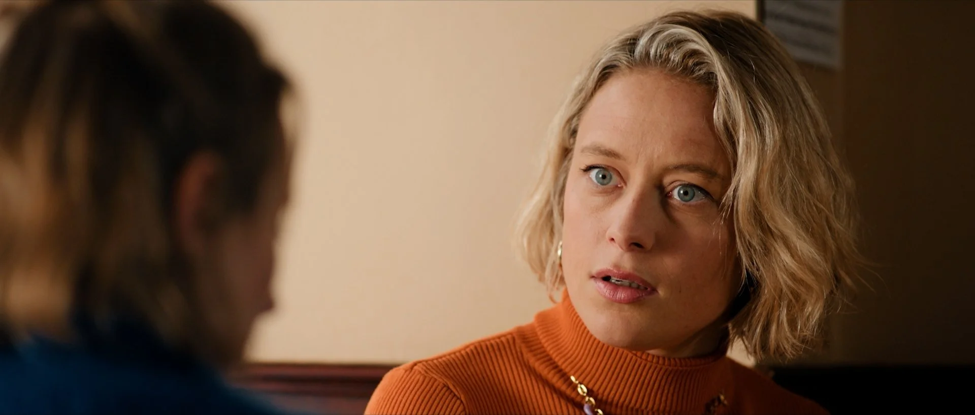 Florence Fauquet with blonde, wavy hair and blue eyes wearing an orange turtleneck sweater and a gold necklace, engaged in conversation with a blurred person.