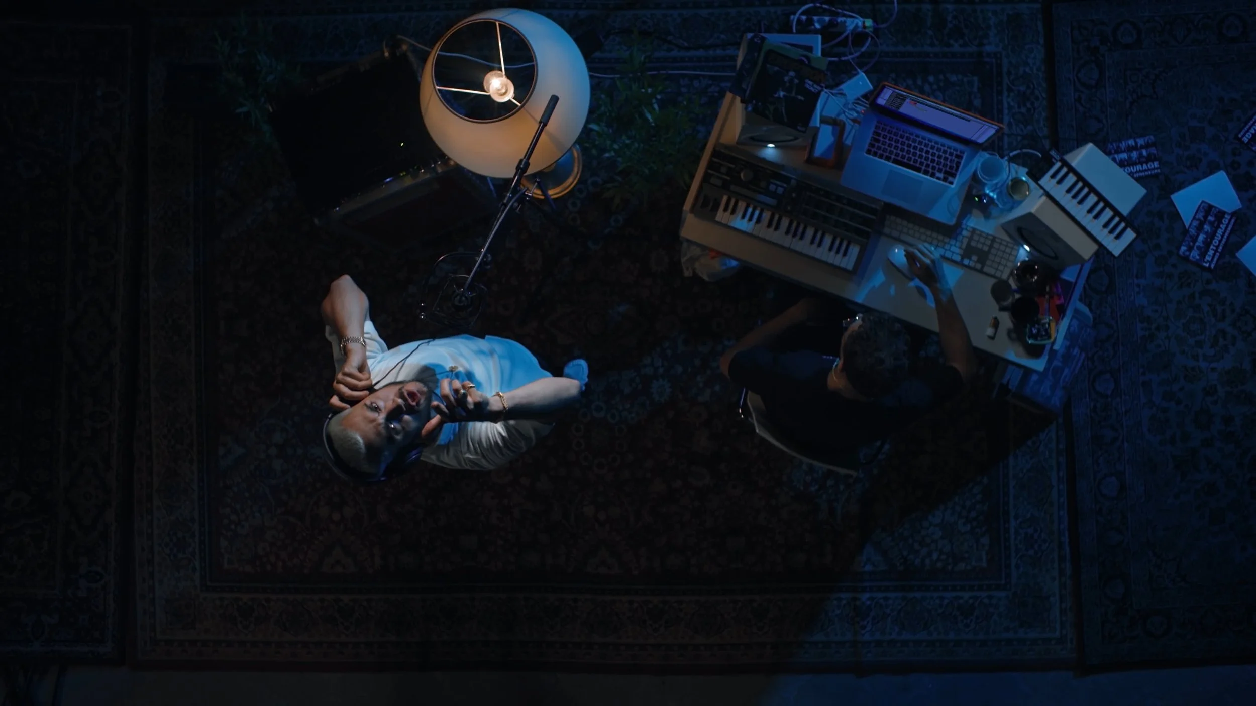 Overhead view of a music studio setup with two people, one singing into a microphone and the other using a computer with music production equipment, including a keyboard and mixing console.
