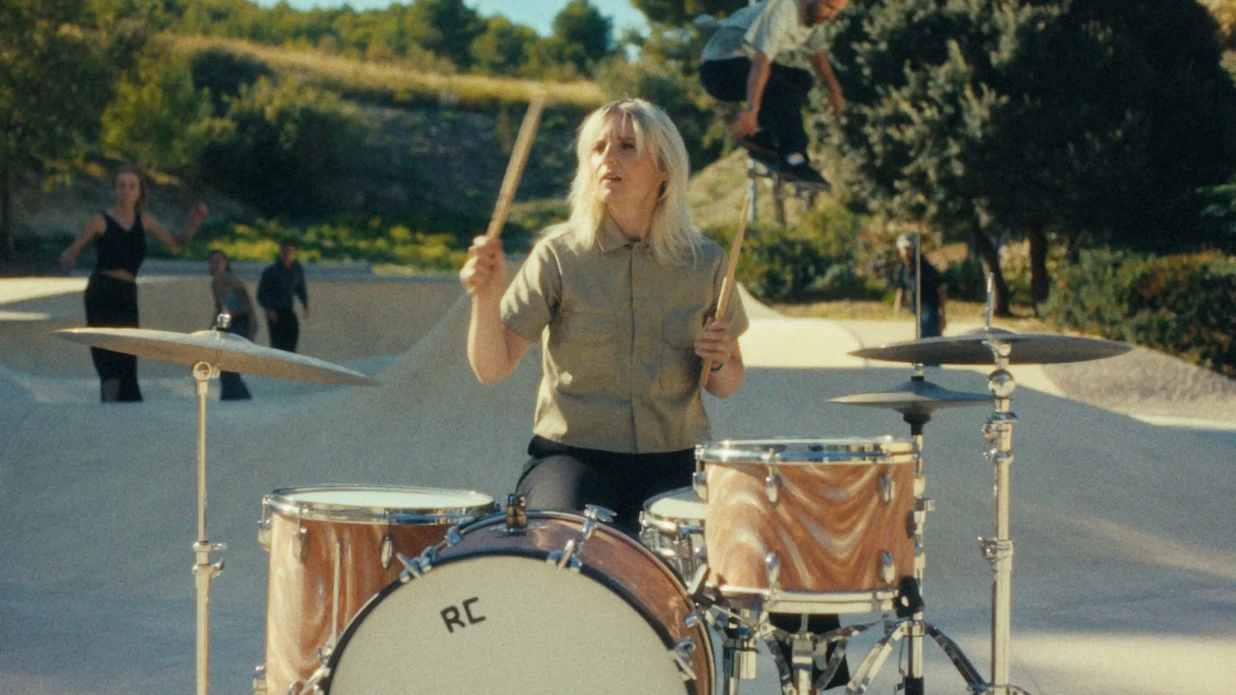 Marion Brunetto from the band Requin Chagrin playing a drum set outdoors in a skatepark while a boy skates in the background, surrounded by trees and greenery.