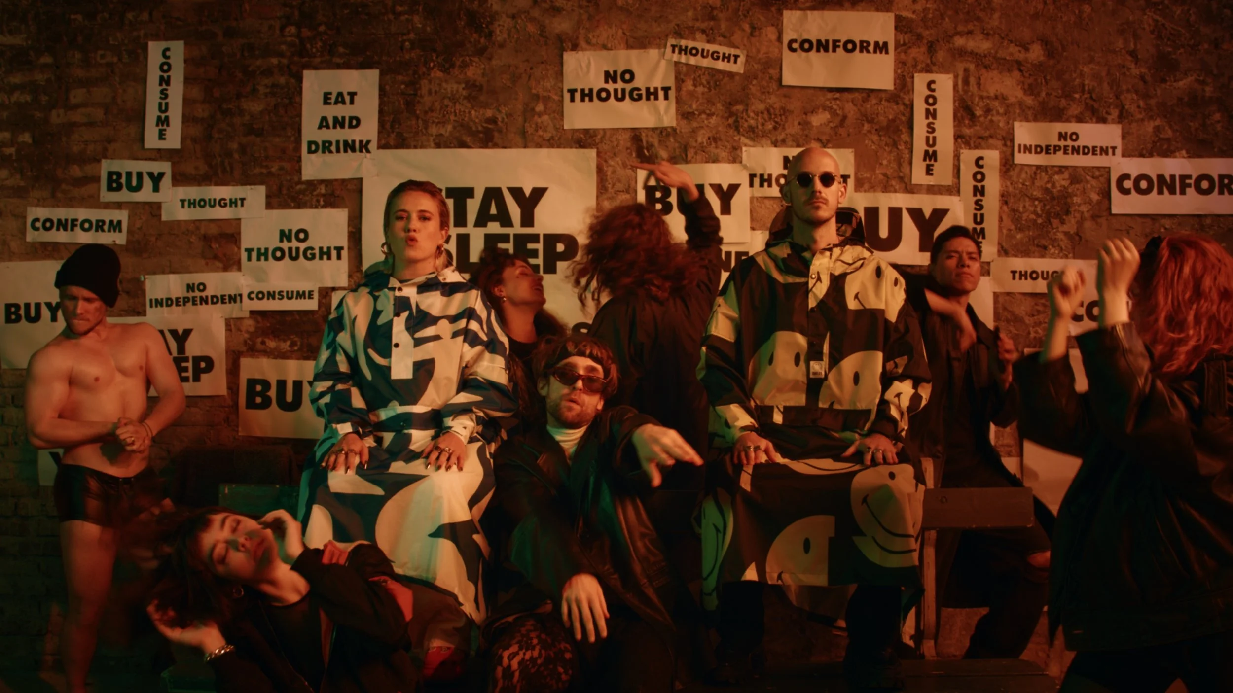 A group of people posing in front of a brick wall with various signs and posters, some with phrases like "STAY SLEEP," "BUY," "CONFORM," and "NO THOUGHT." The individuals display a variety of styles, some dressed casually and others in unique outfits, engaging in different poses and expressions.