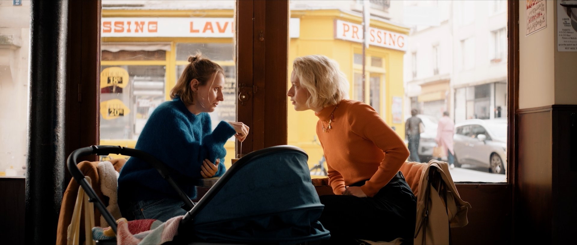 Two women sit face to face in a café near a window, having a serious conversation. Florence Fauquet has blonde hair and wears an orange sweater, while Tiphaine Daviot has brown hair and wears a blue sweater. A stroller is in front of them.