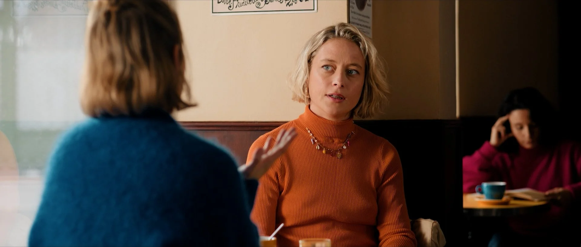 Two women having a conversation in a cafe; Florence Fauquet with short blond hair wearing an orange turtleneck sweater, and the other woman with brown hair wearing a blue sweater. In the background, a woman at a table with a cup and a book.