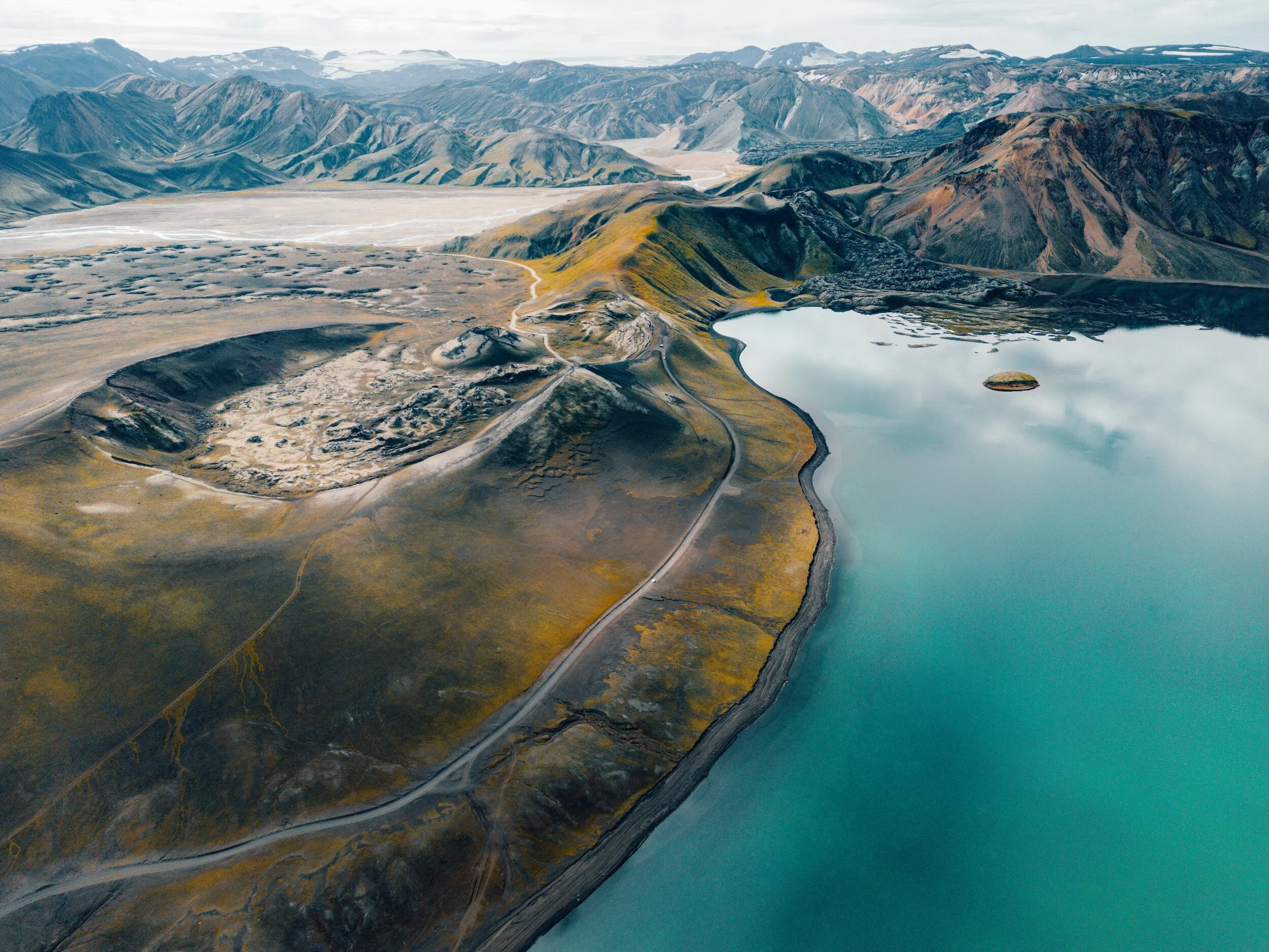Aerial view of a volcanic landscape with a crater, rugged terrain, and a turquoise lake surrounded by mountains.