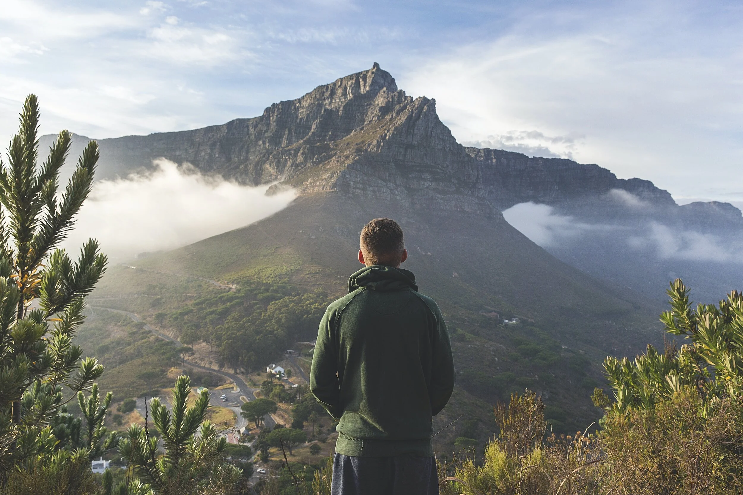 Person in green hoodie overlooking mountain landscape with mist and clouds.