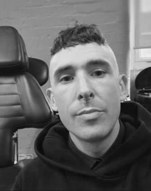 Black and white photo of a young man with short hair, wearing a hoodie, sitting in a room with chairs and a brick wall in the background.