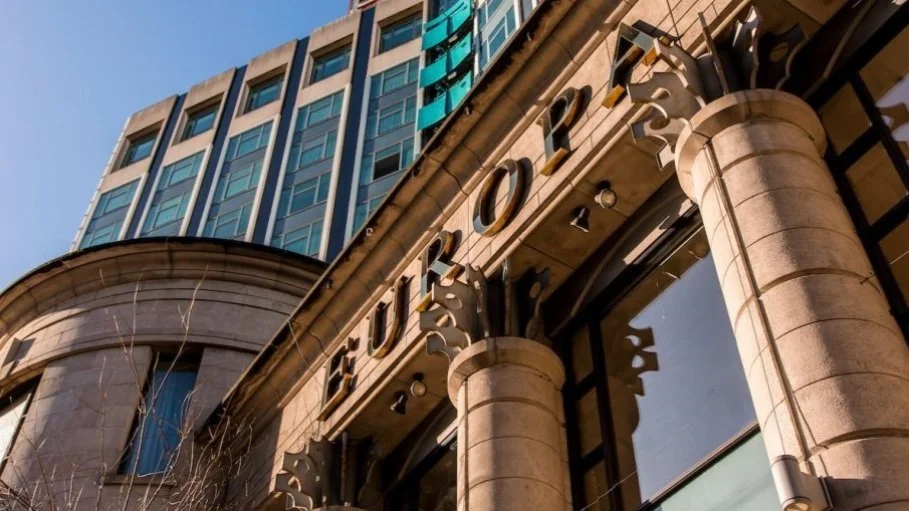 Close-up of a building's first floor showing the sign 'EUROPA' with stone columns and large glass windows, modern upper floors with blue glass windows, part of a city street view under a clear blue sky.