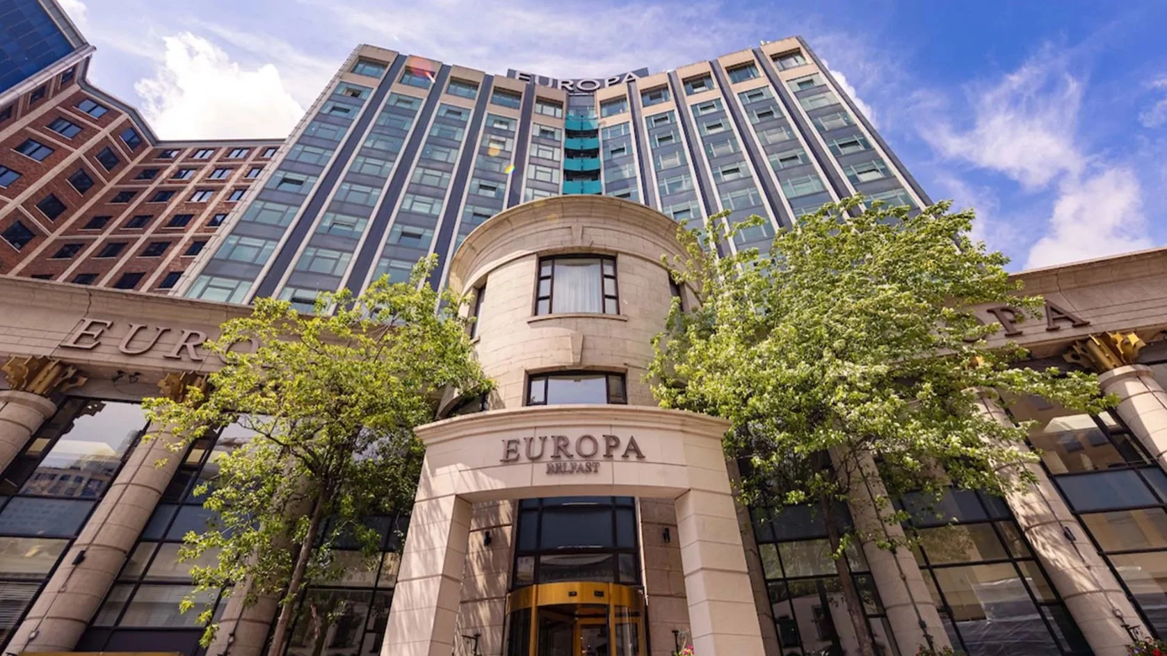 Low-angle view of a tall modern building with signage that reads 'EUROPA' and 'BULAWAY' on the entrance, flanked by green trees, against a partly cloudy blue sky.