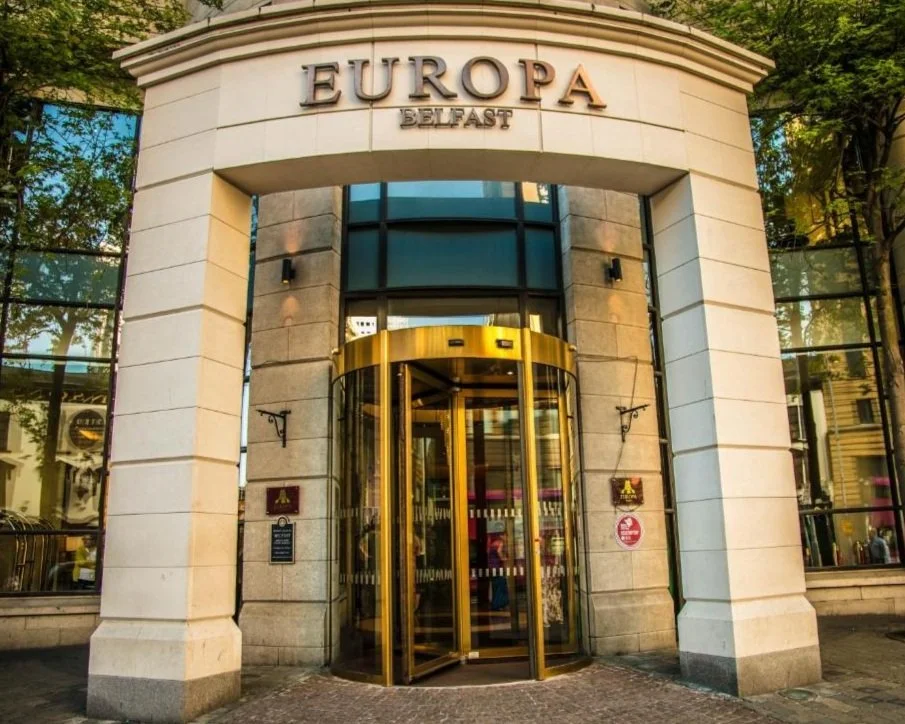 The entrance to Europa Belfast, a building with a rounded glass doorway with a gold-colored revolving door, beige stone walls, and large glass windows reflecting trees and neighboring buildings.