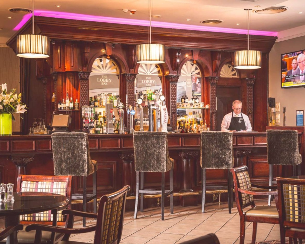 A hotel or pub bar with a wooden counter, bar stools, and taps for drinks. A bartender in a white shirt and black apron is working behind the bar. There are tables and chairs in the foreground, a TV on the wall to the right, and a screen showing a ne